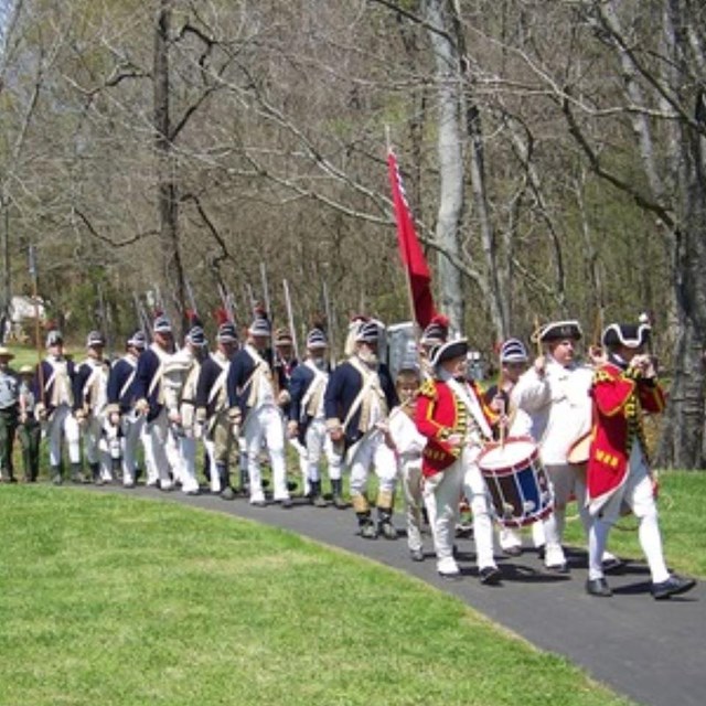 A large group of 18th century dressed soldiers walks along a paved trail
