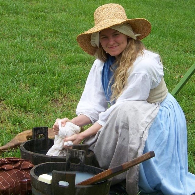 A woman in 18th century clothing is doing laundry with a soapy bucket
