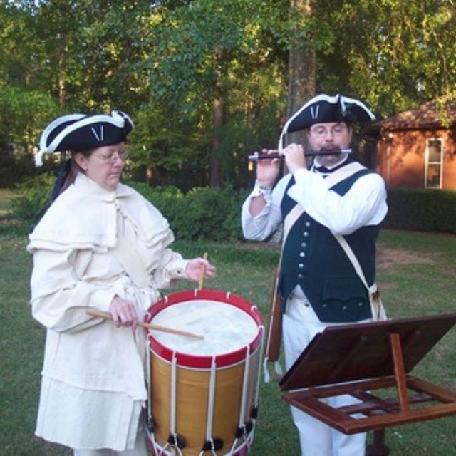 Two people in 18th century clothings are playing a drum and a fife. 
