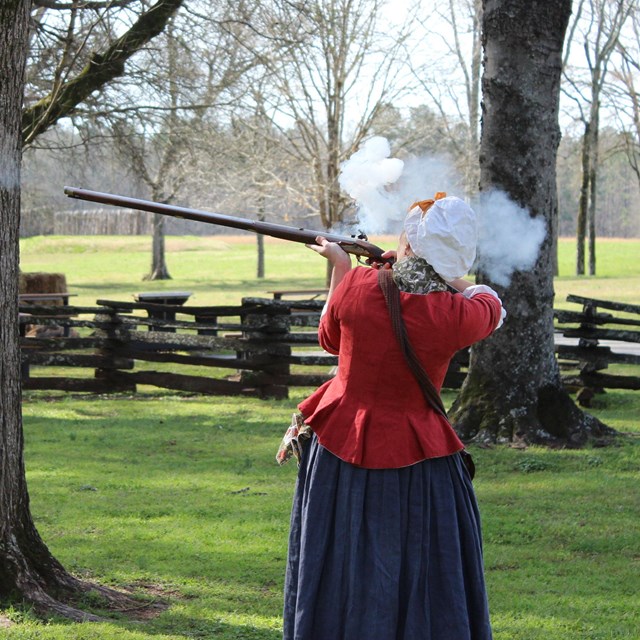 A woman in a red jecket and blue skirt firing a rifle. 