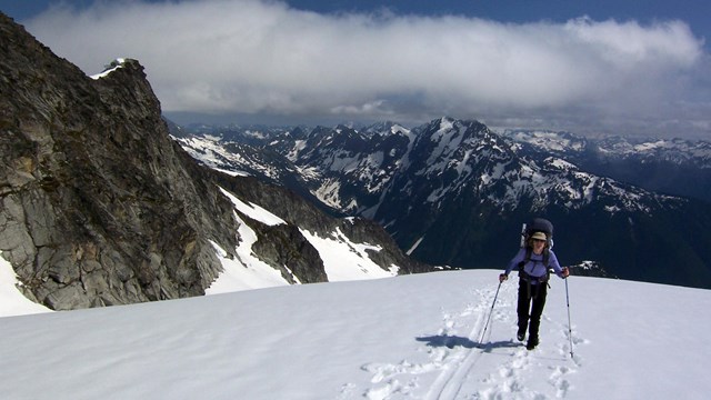 A climber crosses a snow field with mountains in the distance.