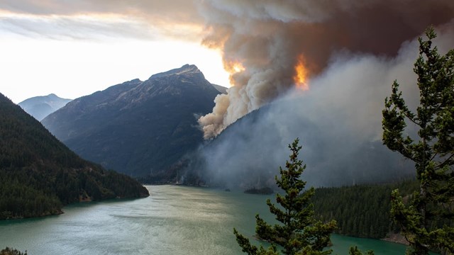 Dark smoke spreading upwards towards the sky next to a mountain with a lake in the foreground