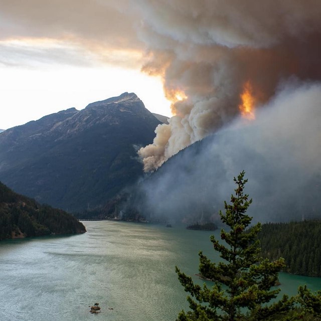 Dark smoke spreading upwards towards the sky next to a mountain with a lake in the foreground