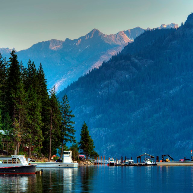 Boats and float plane docked along the shores of Lake Chelan with the mountains rising above. 