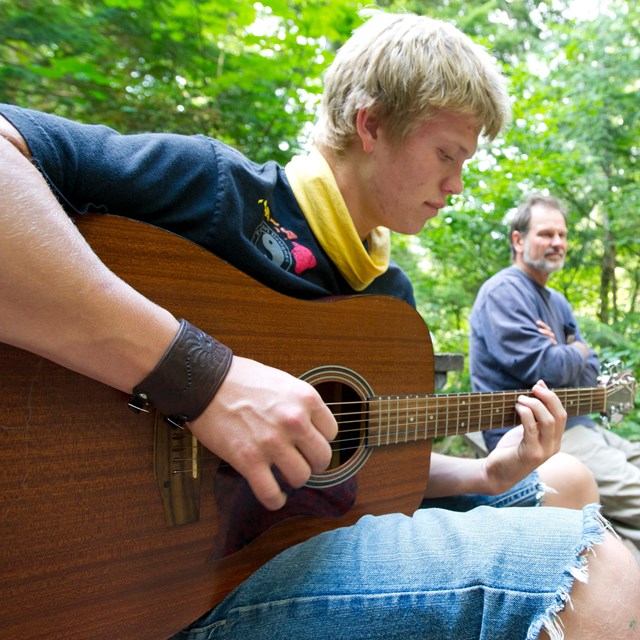 Two men sitting in camp playing guitar 
