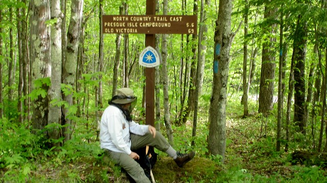 Man wearing a bug net rests along the trail next to a sign.