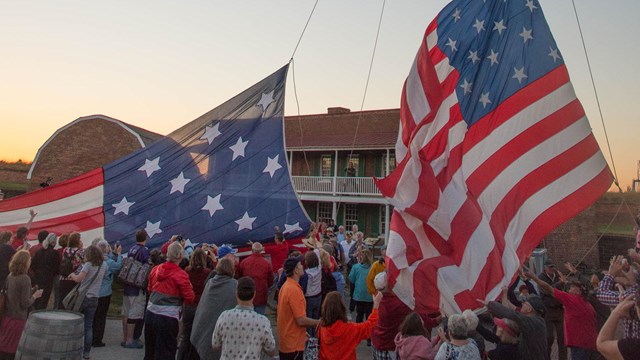 A large group of people help raise two large flags in front of a building.