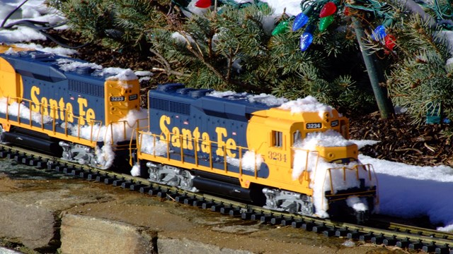 Toy train covered in snow next to evergreen branches with holiday lights