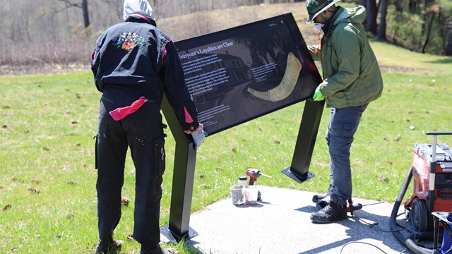 Two people installing a new interpretive sign along a grass-lined walkway.