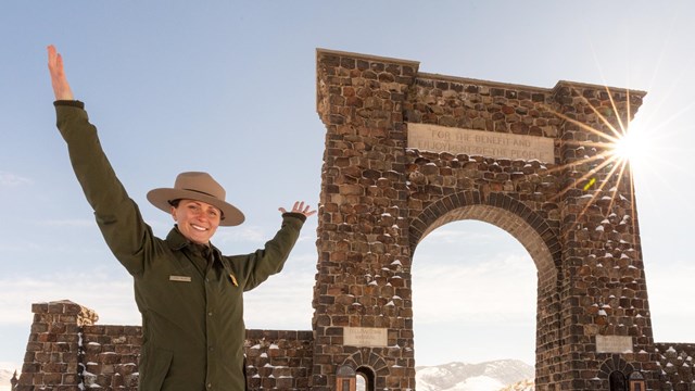 Ranger holding her hands above her head standing in front of the Roosevelt Arch