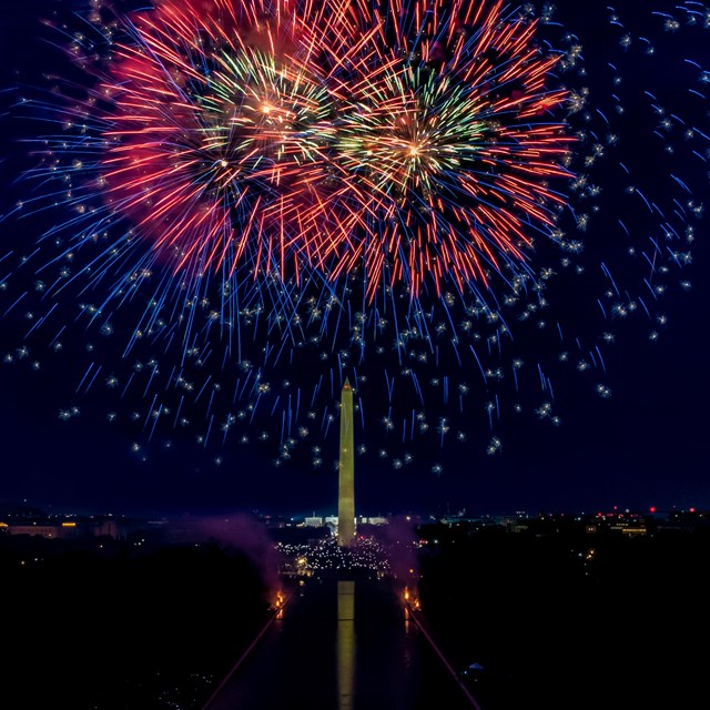 Red and blue fireworks exploding in a dark sky above the Washington Monument and Reflecting Pool
