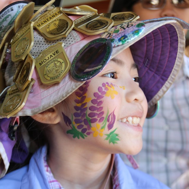 Kid with face paintings wearing a hat covered in Junior Ranger badges