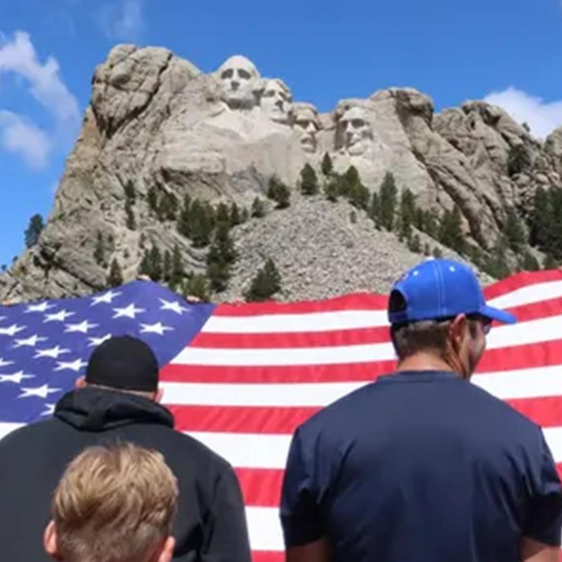 Visitors holding a large U.S. flag under the Mount Rushmore presidents sculpture