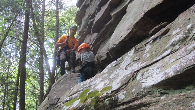 A man and a child climb up a rocky surfaced cliff.
