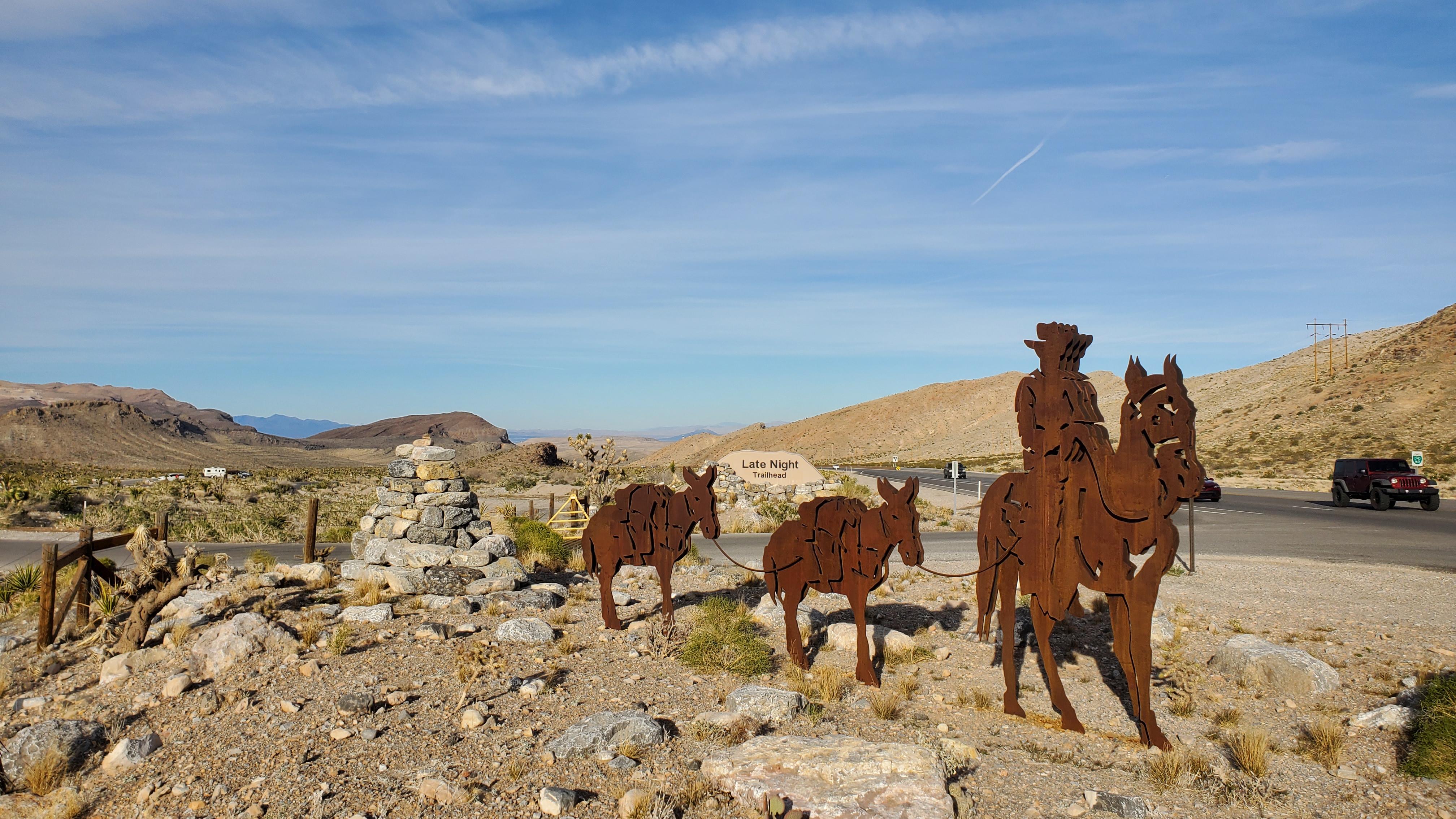Outline statues of a person on burro next to a road in the desert.