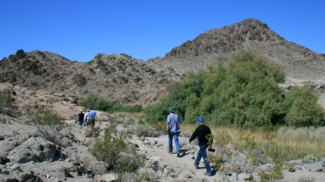 People walk down a trail through a desert landscape with rocky hills and sparse vegetation.