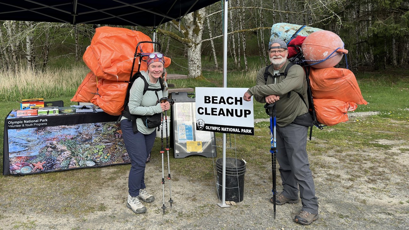 Two volunteers carrying trash from the beach on their back next to a beach cleanup sign.