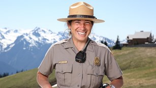 A smiling park ranger wearing a flat hat with mountains in the background.