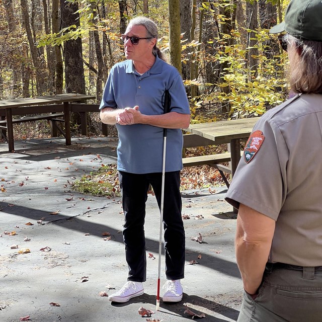 A man wearing sunglasses and using a white-tipped cane stands in a forested area.