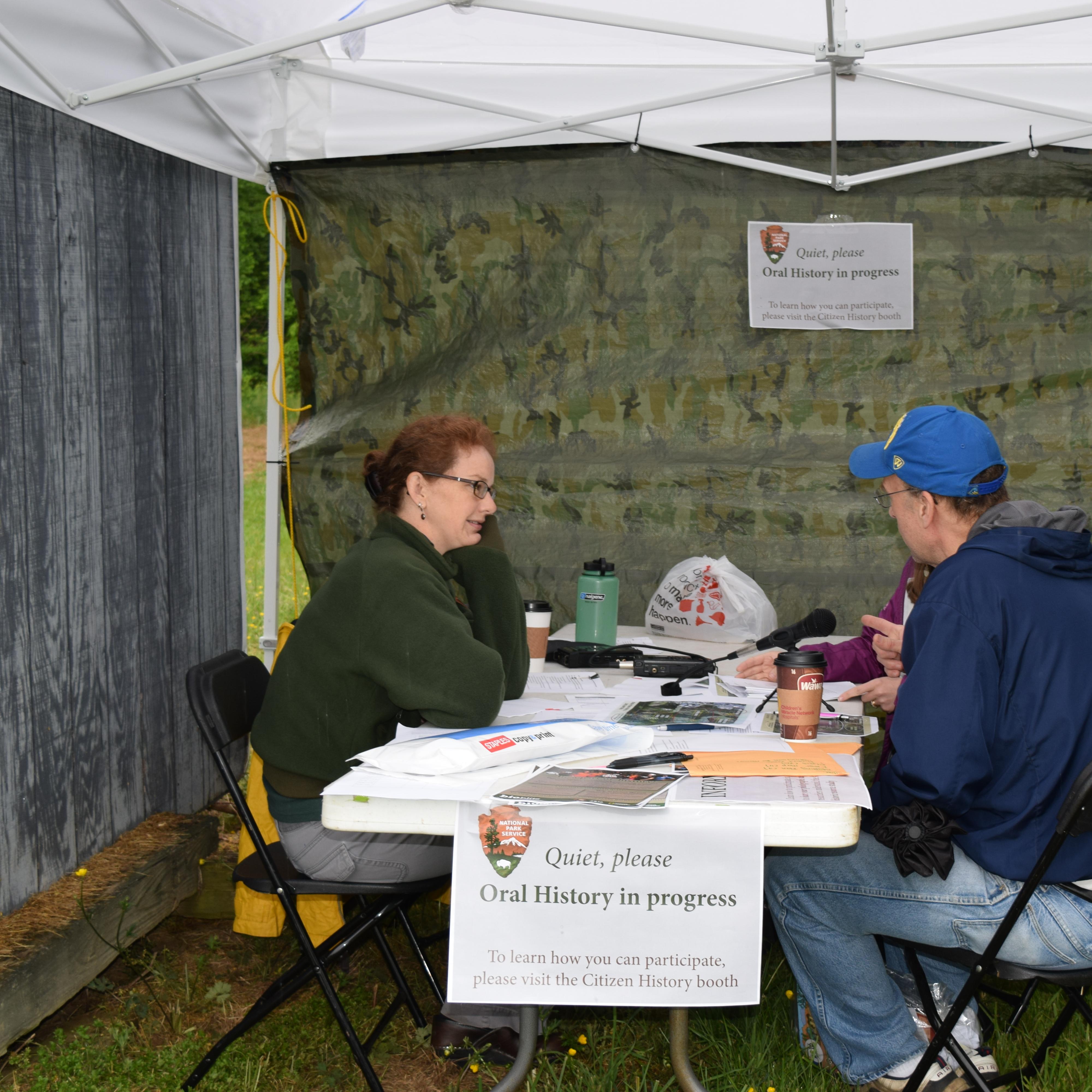 A historian and event visitor sit across from each other at a table under an outdoor tent and talk.
