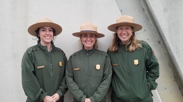 Three uniformed park rangers stand shoulder to shoulder.