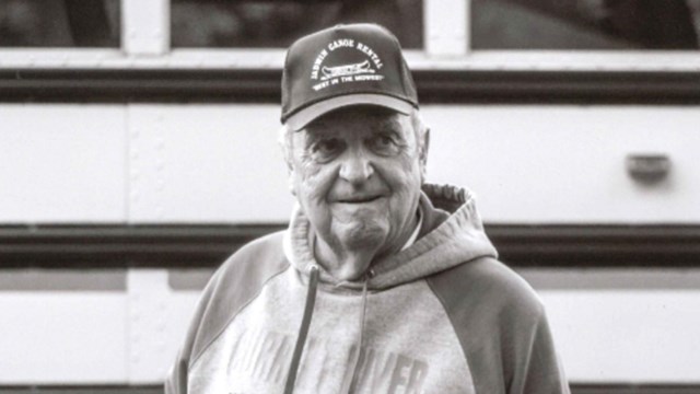 A man wearing a hat and hoodie stands, smiling, in front of a bus.