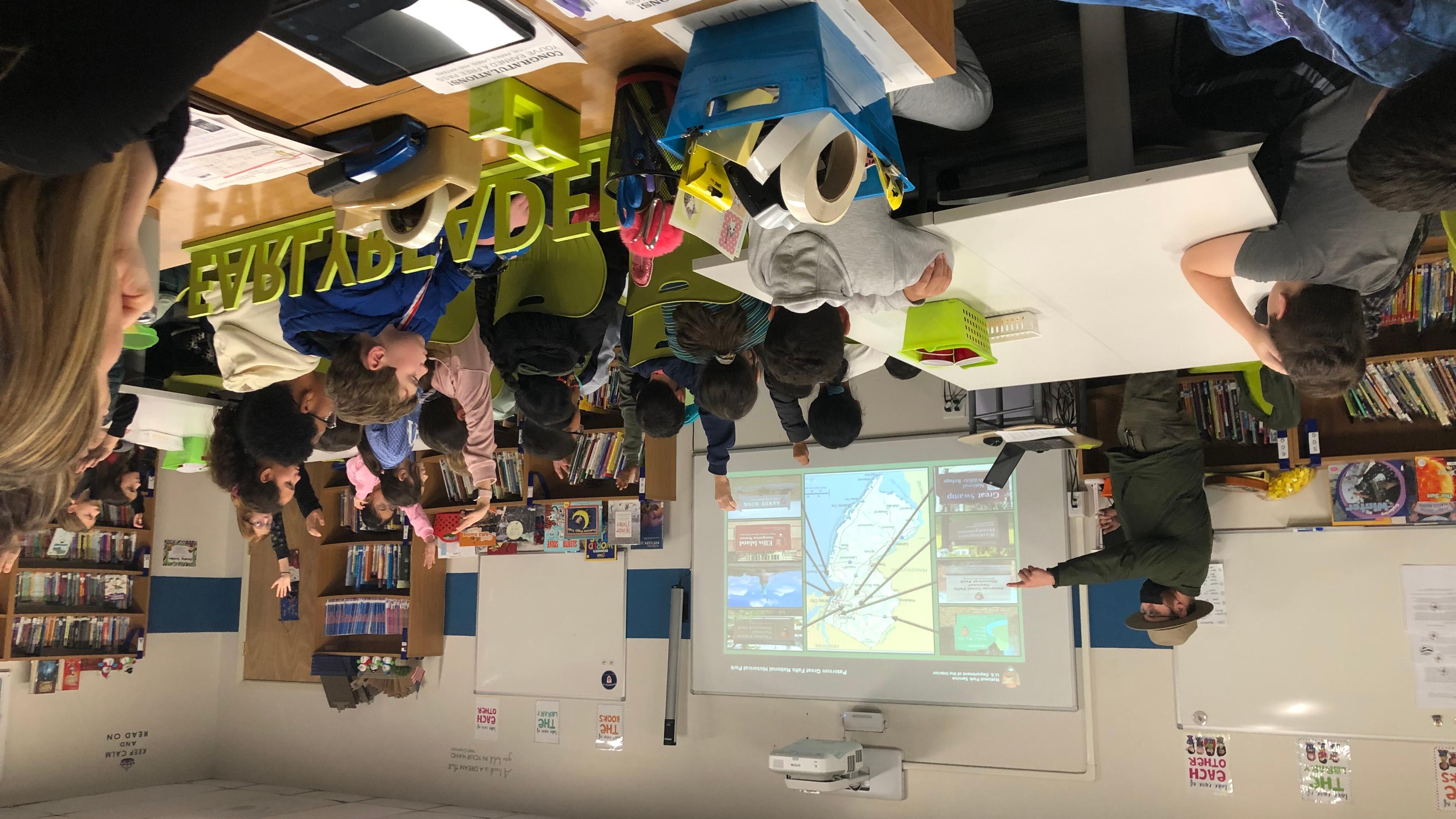 Park ranger pointing as 4th-grade students raise hands in a classroom, a powerpoint on a screen