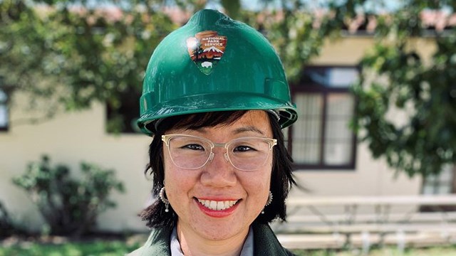 Woman with dark hair, glasses, and green hard hat with NPS logo stands in front of white building