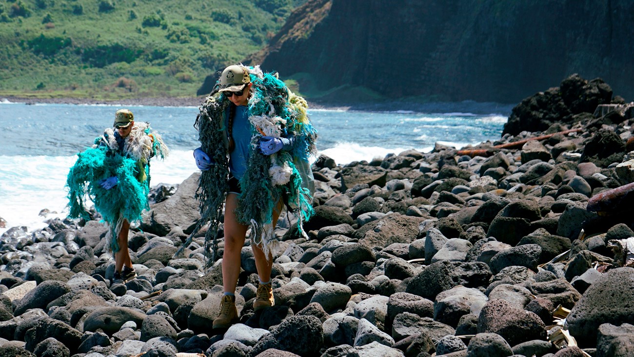 Two people carrying nets and beach debris, walking on a cobbled beach 