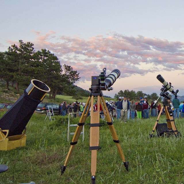 A group of people with telescopes standing in a grassy field at sunset