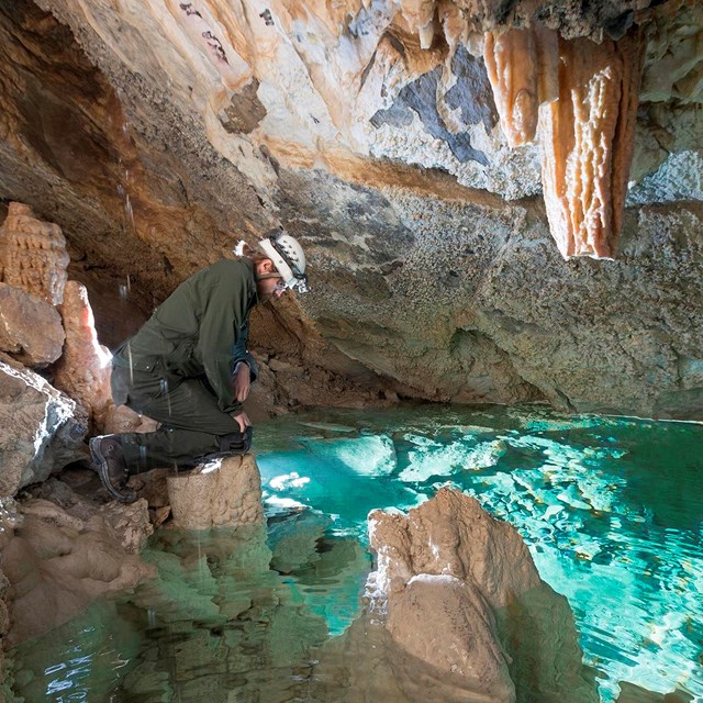 A man wearing a hard hat kneels next to a turquoise lake surrounded by cave formations