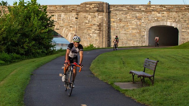 Group of bicyclists on a paved trail leading from a stone bridge.