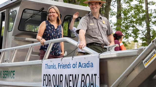 Two people stand on a new boat with a banner reading "Thanks, Friends of Acadia FOR OUR NEW BOAT."