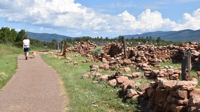 A woman with a dog walks next to the low rock walls of Pecos Pueblo