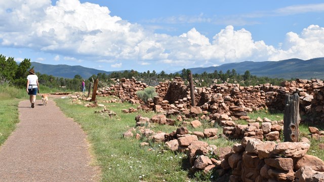 A woman with a dog on a trail next to the low walls of Pecos Pueblo