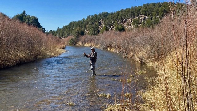 An angler stands in the Pecos River, red and tan brush lining the river