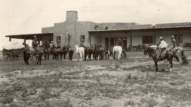 Historic photo of a herd of horses, three men mounted, chuckwagon on left, long covered porch.