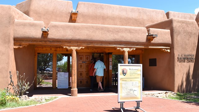 Front of main visitor center, a tan adobe building, with two visitors entering