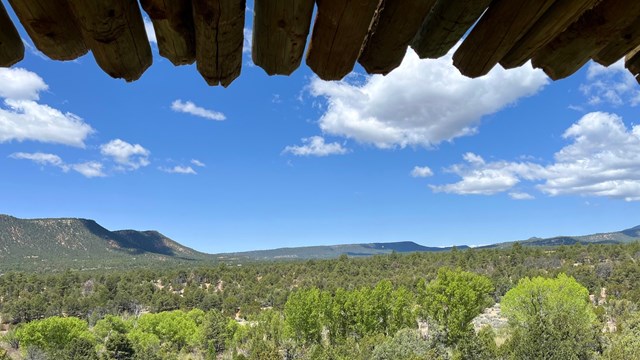 Ramada roof beams extend over the top of the view toward green Glorieta Mesa and Gloria Pass.