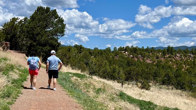 Two people walk a gentle trail with blue sky, trees and light clouds.