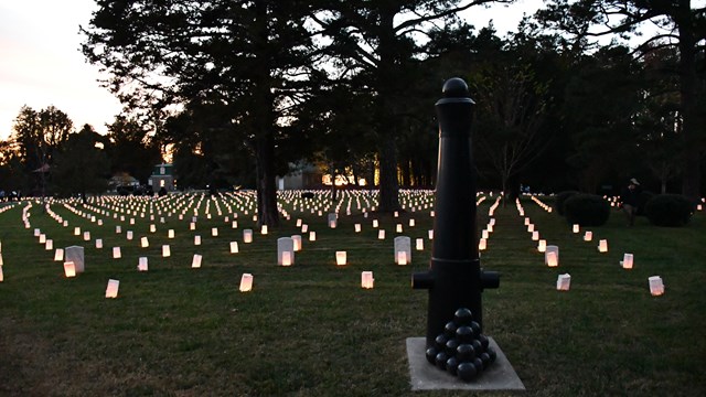 A pyramid of cannon balls in front of cannon tube in its end with luminaries in background.