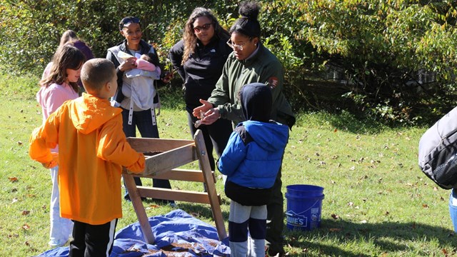 Three children and two adults listen to a female in NPS uniform talk about an archeology sifter.