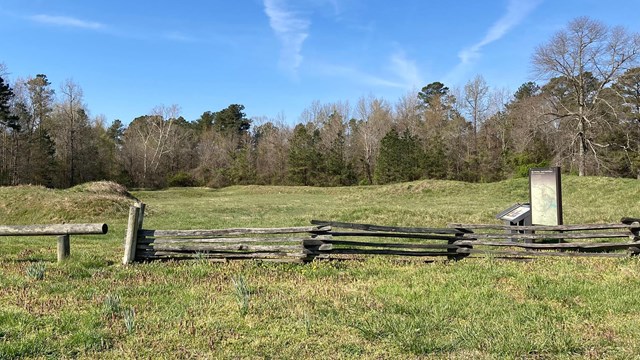 A split rail fence in front of green grass covered earthworks.