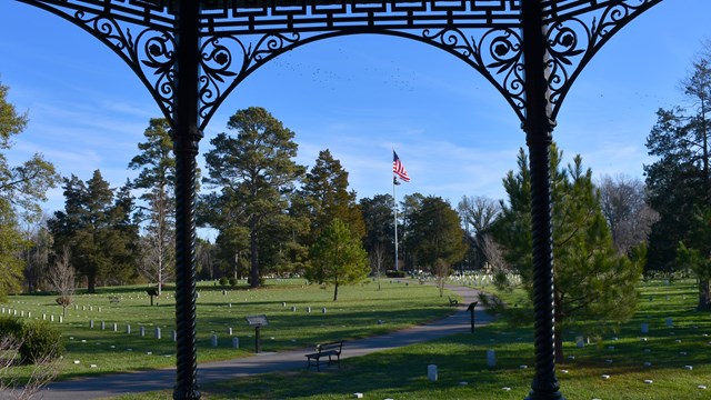 Wrought iron columns frame a view of white headstones surrounded by green grass.   