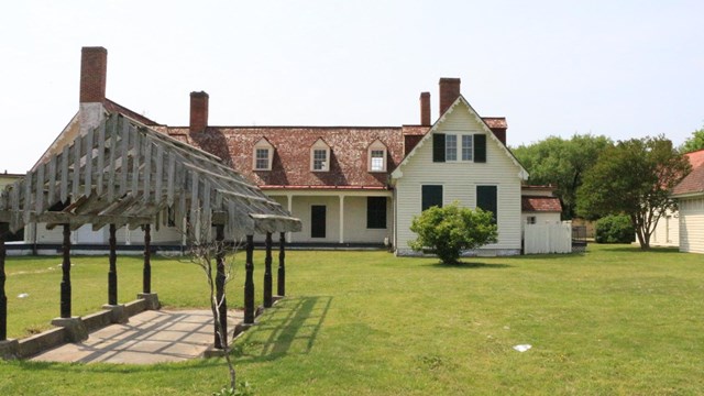 An arbor on left and a two-story white building with red roof in the background and a green lawn.