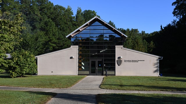 A wide sidewalk leads to barn shaped tan building the center section is glass windows.