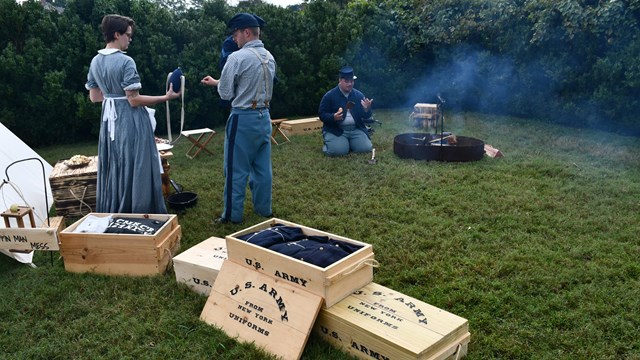 Three living historians cook breakfast at a fire pit with boxes of supplies in the foreground. 