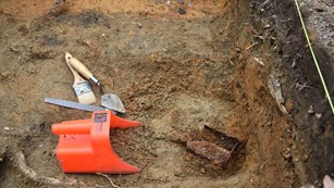 Bucket, brush, and trowel sit on ground in a hole next to an artifact.