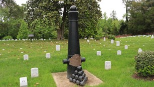 Pyramid of cannonballs in front of a cannon tube in front of headstones. 
