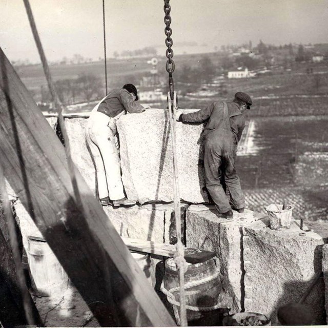 a black and white photo of 2 men standing on stacked blocks of granite while receiving another block